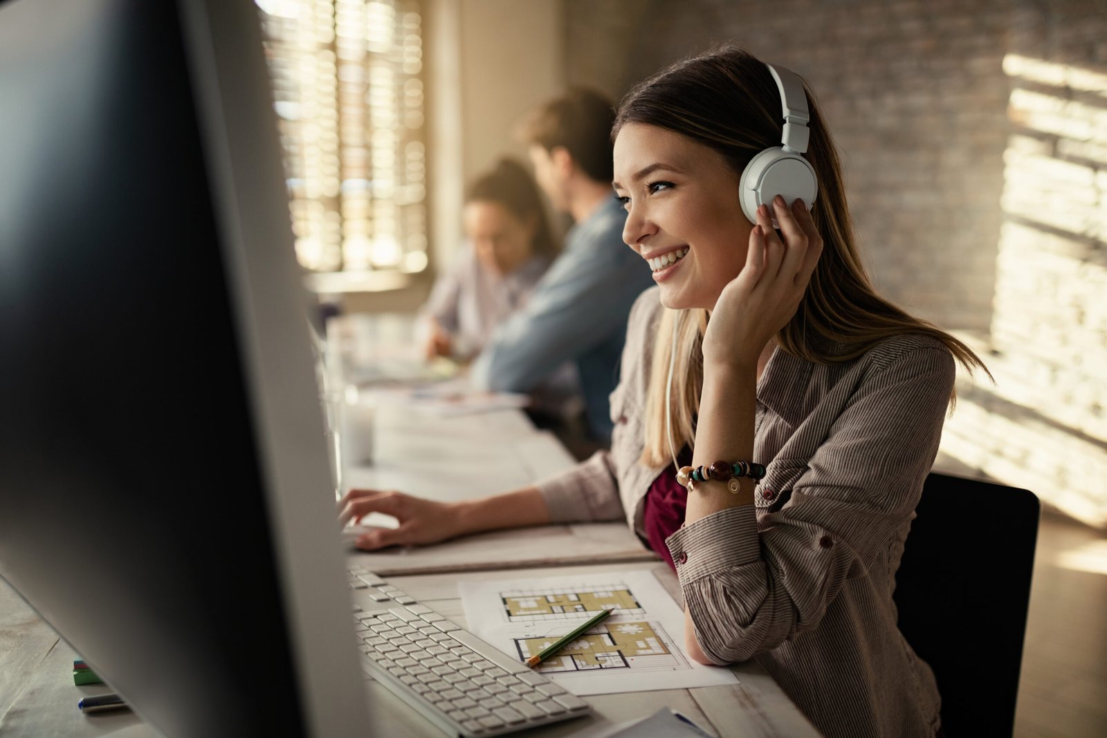 Young happy businesswoman working on a computer and listening music over headphones in the office. Her colleagues are working in the background.